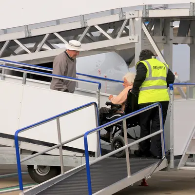 Airport security staff helping an elderly person in a wheelchair as they prepare to board or disembark from an aircraft. A companion or assistant is standing nearby, ready to support. The wheelchair is positioned at the bottom of a portable ramp leading up to the plane, ensuring safe and accessible mobility for the passenger. The setting emphasizes airport assistance and accessibility services for travellers with mobility challenges.