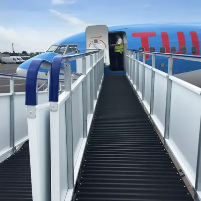 Aircraft boarding bridge connected to an airplane, allowing passengers to board or disembark safely. The bridge is made of metal and has a walkway with a non-slip surface. An airport staff member is standing near the entrance, possibly assisting passengers. The setting is on the airport tarmac under a clear sky, showcasing ground support equipment used in aircraft operations.