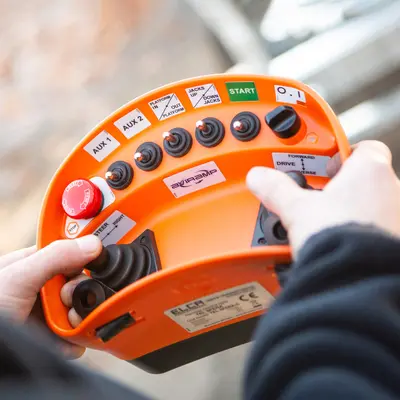 A professional holding a sophisticated remote control device, possibly used for operating a drone, remote-controlled vehicle, or industrial equipment. The remote has multiple buttons, switches, and joysticks, suggesting it is designed for precise and versatile control. The person’s hands are firmly gripping the device, indicating they are actively operating or preparing to operate the equipment. This setup is commonly seen in activities such as drone piloting, remote machinery operation, or remote-controlled hobbyist vehicles, emphasizing the importance of careful handling and coordination.