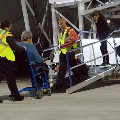 Airport staff helping a passenger in a wheelchair as they use a small mobile staircase to board or leave the aircraft. The staff members are wearing safety vests and are assisting with care and support. The setting is on the airport tarmac, showing ground support activities focused on passenger assistance, especially for those with mobility challenges. Overall, it highlights the airport's efforts to provide accessible and helpful services for all travellers.