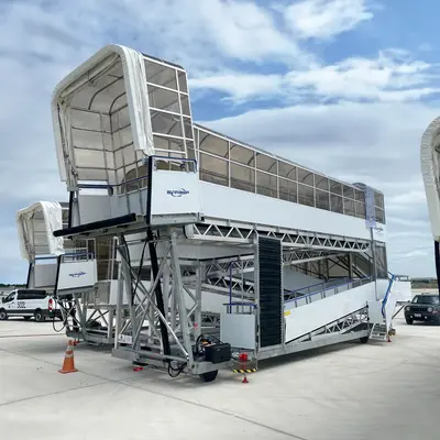 An airport passenger staircase or mobile jet bridge, designed to connect the ground to an aircraft for boarding and deplaning. It's a portable structure with multiple levels, supported by a sturdy framework, and positioned on the tarmac. The setup includes safety features like railings and cones, and the sky suggests it's daytime. Overall, it shows a typical piece of ground support equipment used at airports to assist passenger movement when fixed jet bridges aren't available.
