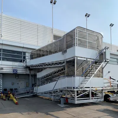 A mobile passenger stairway at an airport, connected to an aircraft or positioned ready for use. The stairway allows passengers to board or leave the plane safely and efficiently. It has multiple steps and safety railings, and is designed to be movable for easy positioning. The background suggests an airport environment, with the terminal building nearby. Overall, it shows a typical airport ground support equipment used during aircraft turnaround operations.