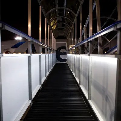 A modern, enclosed pedestrian walkway or footbridge, probably at an airport or transportation hub. It is well-lit with sleek lighting, and the transparent sides allow travelers to see outside as they walk through. The curved roof and clean design give it a futuristic and safe appearance, guiding pedestrians smoothly toward their destination. Overall, it’s a contemporary structure built for passenger convenience and safety.
