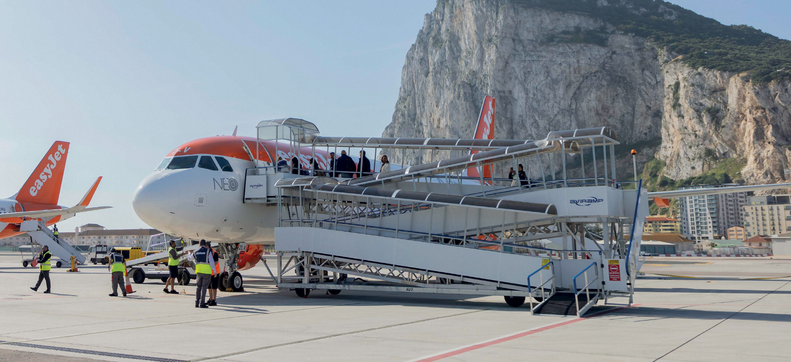 A vibrant airport scene featuring a bright, modern easyJet airplane parked on the tarmac. The aircraft is positioned next to a mobile passenger staircase, which allows travelers to board or leave the plane conveniently. The background showcases a stunning mountainous landscape under a clear, sunny sky, adding a dramatic and scenic element to the scene. Around the aircraft, airport staff and crew are busy with ground operations, ensuring everything runs smoothly. The overall atmosphere is lively and efficient, highlighting the dynamic environment of air travel, all set against a striking natural backdrop that makes this scene truly captivating!