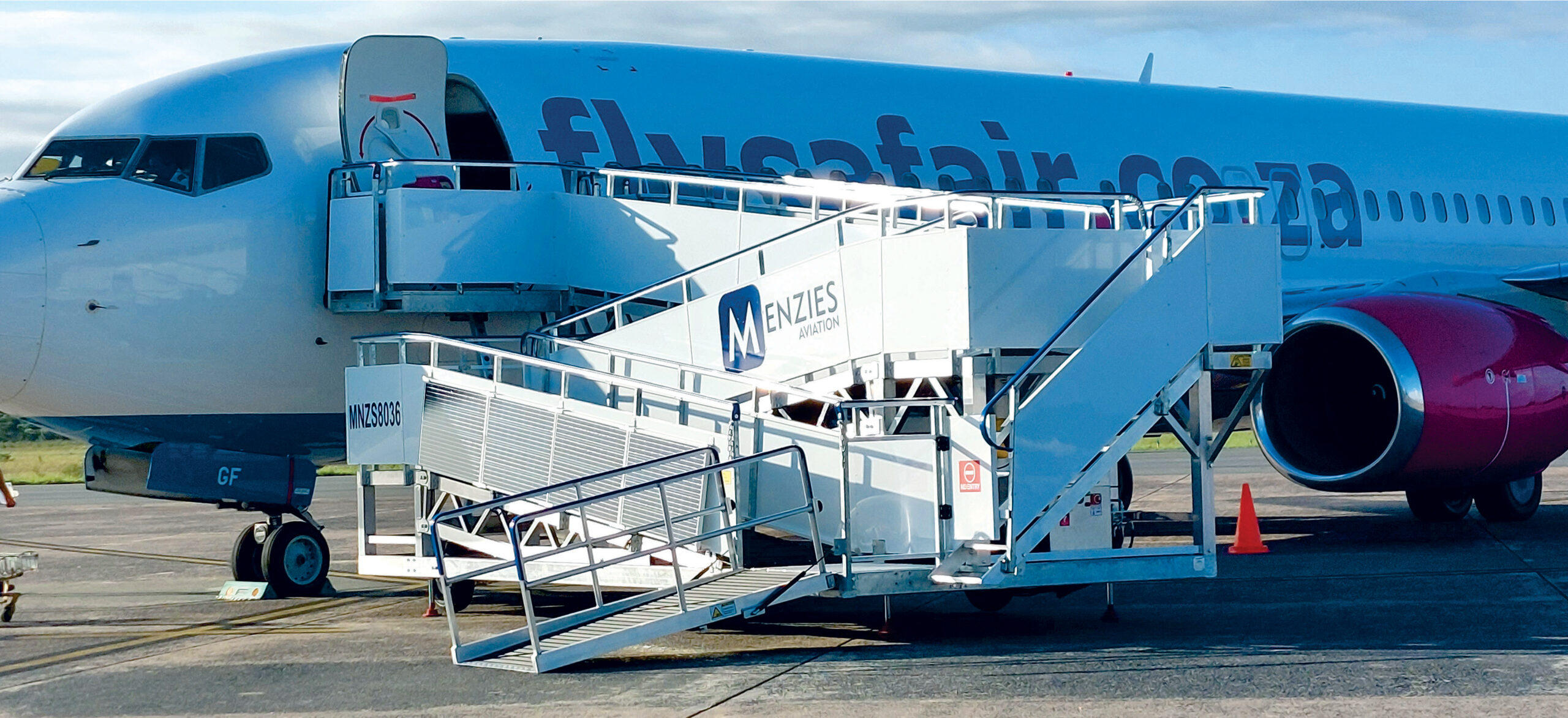 One aircraft parked at the airport with a mobile boarding staircase attached, allowing passengers to board or disembark. The aircraft is serviced by ground handling company Menzies Aviation, as indicated by the branding on the stairs. The scene is set during daylight hours with clear weather, making it an ideal day for travel. This setup is typical for airports that use portable stairs instead of jet bridges, especially at remote stands or smaller airports. The overall atmosphere suggests efficient ground operations and smooth passenger movement.
