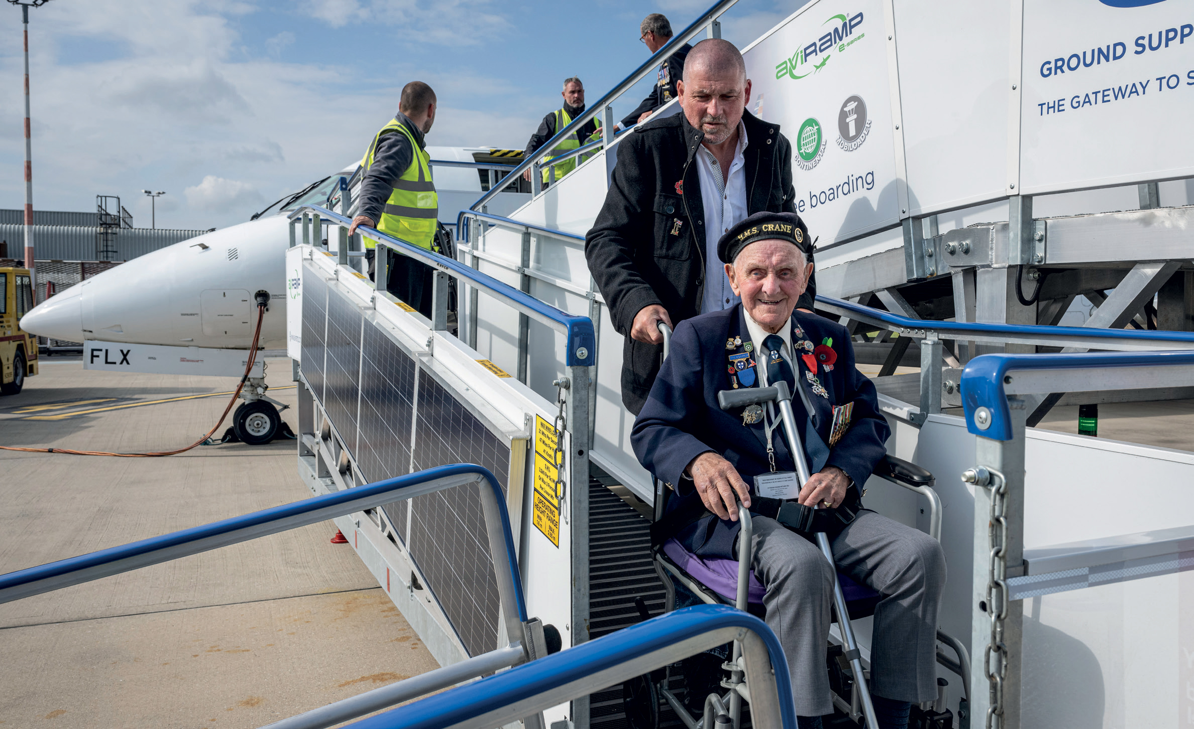 This touching scene captures a veteran being assisted as he disembarks from an aircraft, likely during a special flight or event honoring his service. Dressed in a formal military uniform decorated with medals and ribbons, he exudes pride and dignity. The veteran is seated in a wheelchair, emphasizing the importance of accessibility and care during travel. A kind companion or handler is helping him down the portable steps, ensuring his safety and comfort. The setting appears to be an airport tarmac, with ground crew and equipment nearby, hinting at a well-organized, respectful occasion—possibly a veterans’ flight, a commemorative event, or a special return home. The background shows a clear sky and airport infrastructure, adding to the atmosphere of a proud and heartfelt tribute. This scene beautifully highlights the respect and gratitude extended to those who served, celebrating their bravery and sacrifice with dignity and honour.