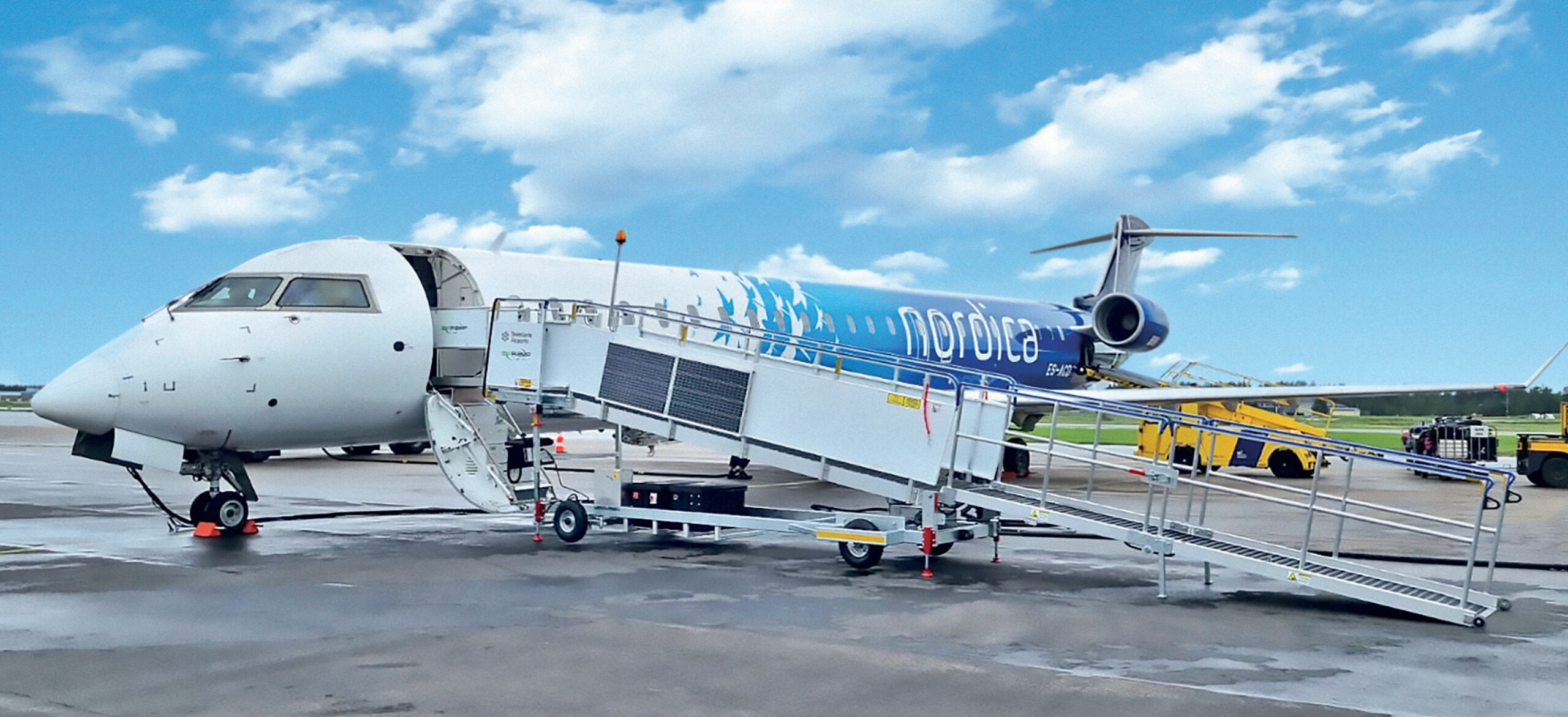An airplane parked at an airport on a beautiful, clear day with bright blue skies and minimal clouds. The aircraft appears to be ready for boarding or disembarking, connected to a portable passenger staircase. The scene suggests calm weather, ideal for travel, and highlights typical airport operations. The blue sky adds a serene and inviting atmosphere, making it look like a pleasant day for flying.