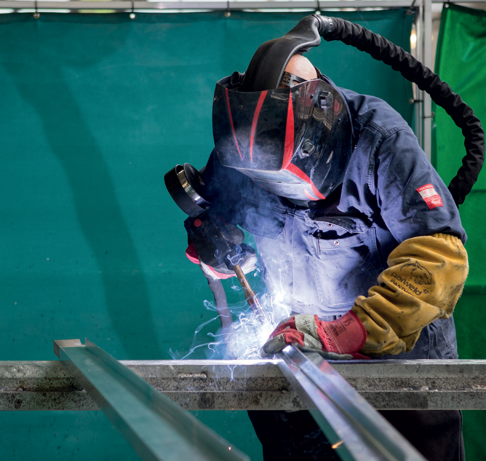 Vividly illustrating the art and science of welding, a crucial process in construction, manufacturing, and metal fabrication. The welder’s protective gear, including the helmet with a darkened visor, gloves, and flame-resistant clothing, underscores the importance of safety when working with intense heat and bright sparks. The glowing sparks and smoke highlight the high energy and precision involved in melting and fusing metal pieces together. The background, with its green curtain or partition, suggests an industrial workshop or a controlled environment designed to contain sparks and protect other workers from hazards. The focus and concentration of the welder reflect the skill required to execute precise welds, ensuring the strength and durability of the finished product.