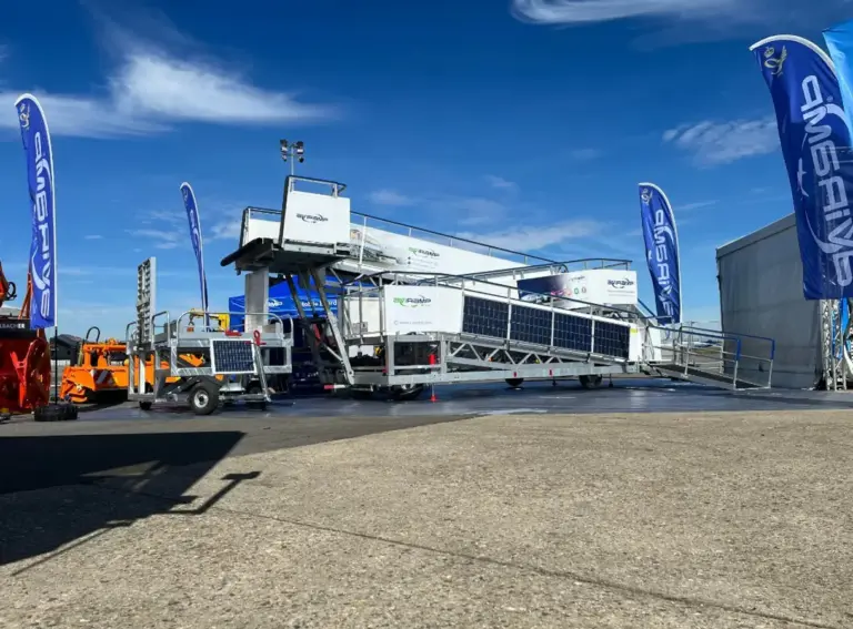 A large portable aircraft boarding ramp positioned outdoors at an aviation event, with flags and equipment nearby under a clear blue sky.