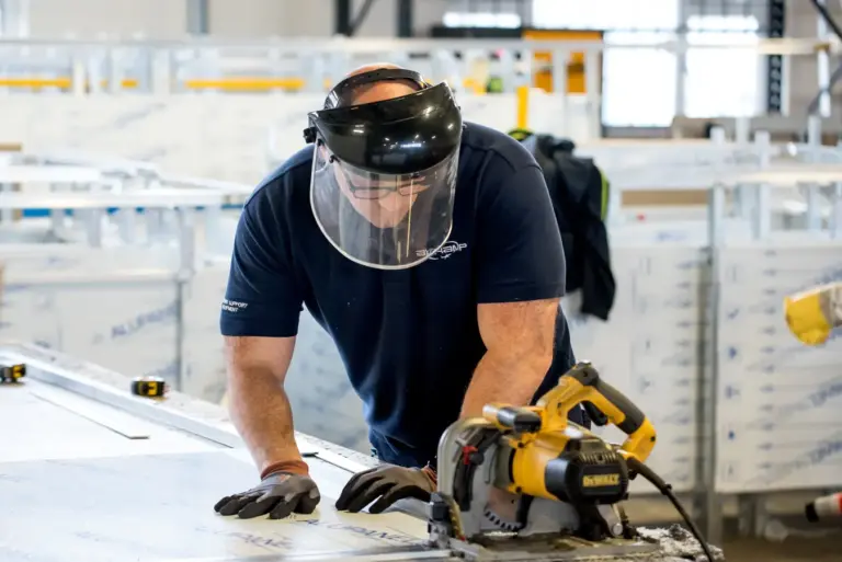 A muscular man engaged in a demanding task within an industrial or construction setting. His well-built physique suggests he is accustomed to physically intense work, emphasizing both power and endurance. Wearing essential safety equipment—such as gloves and a face shield—he demonstrates a commitment to safety while handling heavy-duty tools like a power saw. The focus and precision on his face indicate his experience and concentration, ensuring the task is completed efficiently and safely. The industrial backdrop, with tools and materials around him, underscores the environment where strength and technical skill are vital. This image celebrates craftsmanship, physical fitness, and the importance of safety protocols in manual labour jobs. Overall, it portrays a hardworking individual using his strength and expertise to accomplish a challenging task, embodying resilience, skill, and dedication in a tough working environment.
