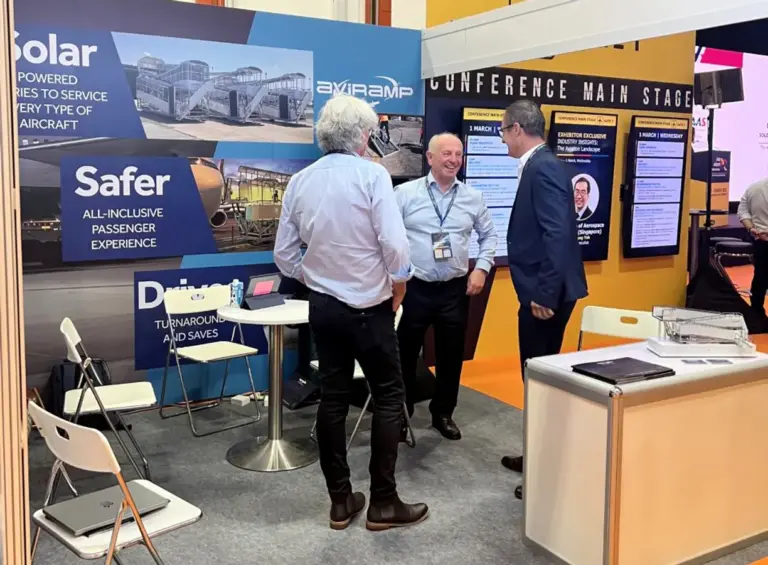 Three men having an animated conversation at an aviation trade show booth, surrounded by displays and seating, likely discussing products or industry topics.