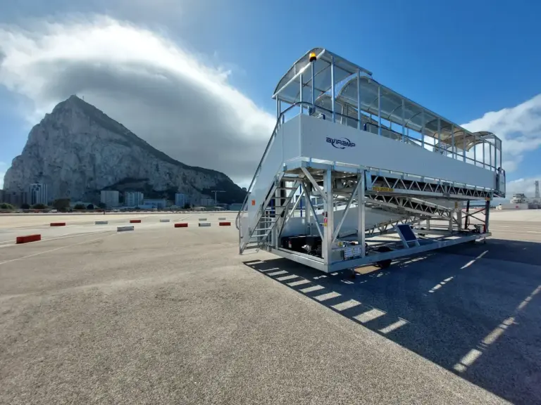 A key moment at the airport, where a mobile boarding staircase stands ready beside an aircraft, facilitating the movement of passengers from the terminal to the plane. The presence of the iconic Rock of Gibraltar in the background adds a distinctive and recognizable landmark, emphasizing the location's unique geographical and cultural significance. The partly cloudy sky and sunlight create a bright, inviting atmosphere, evoking feelings of anticipation and adventure associated with travel. The scene showcases the seamless coordination involved in airline operations—ensuring passengers move safely and efficiently between the ground and aircraft. The backdrop of Gibraltar’s famous rock not only provides a stunning visual element but also hints at the importance of this location as a gateway between continents, symbolizing connectivity and exploration. Overall, this image encapsulates the excitement of embarking on a journey, set against the striking scenery of a historic and vibrant destination.