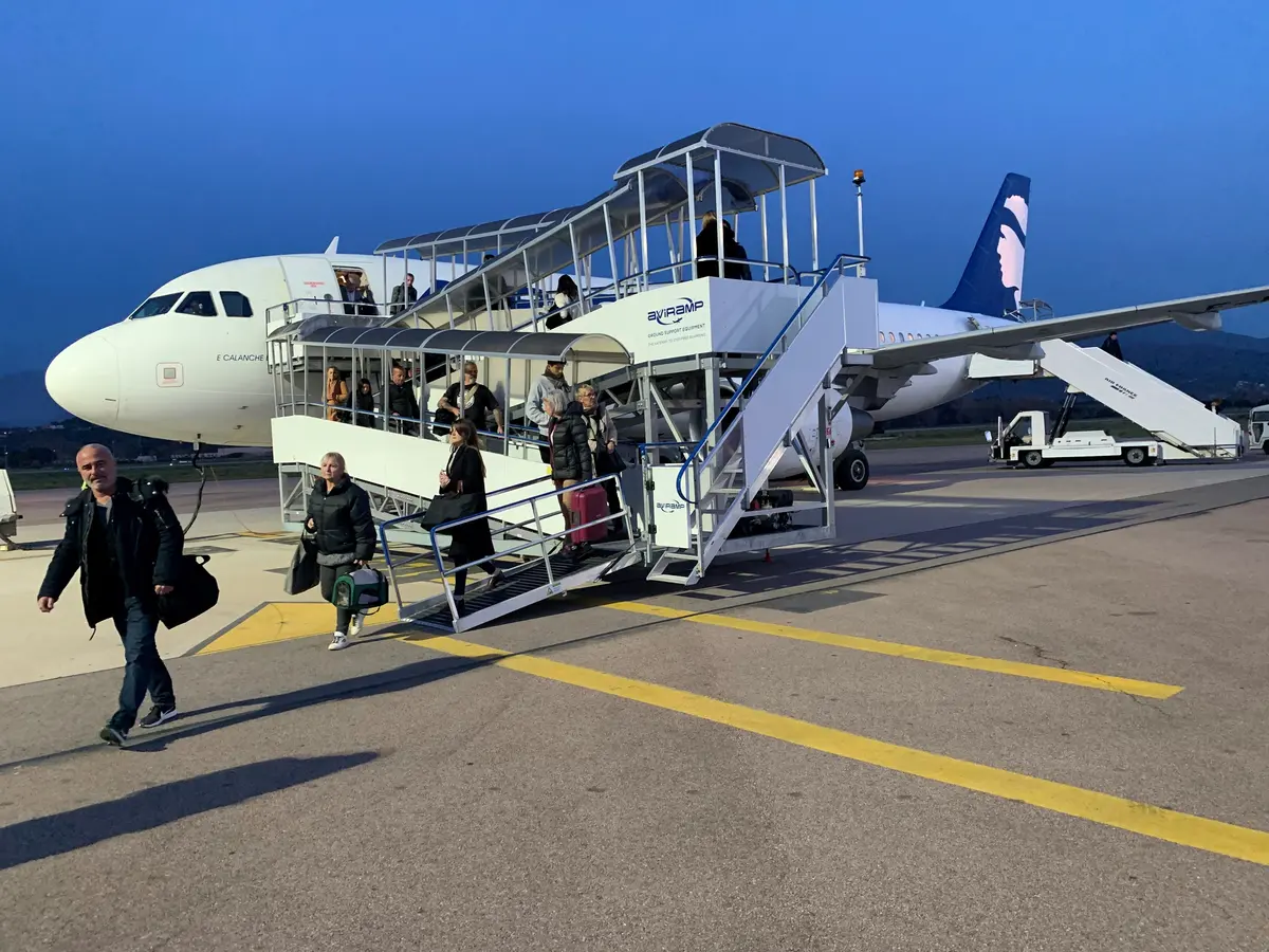 A bustling airport moment during the evening hours, where travelers are actively boarding a commercial aircraft using a mobile staircase. The soft glow of the setting sun or evening lighting creates a warm and inviting atmosphere, highlighting the vibrancy of travel even as the day winds down. Passengers carrying their luggage or personal belongings are making their way up the stairs, indicating a mix of excitement and anticipation for their journey ahead. Airport staff and crew members are present, providing assistance to ensure a smooth boarding process, reflecting the professionalism and hospitality inherent in air travel. The background, with the evening sky transitioning into twilight, adds a sense of calm and serenity, emphasizing the global connectivity that air travel provides. Overall, this scene embodies the dynamic energy of airports—where stories begin, adventures unfold, and the world becomes more accessible with each flight.