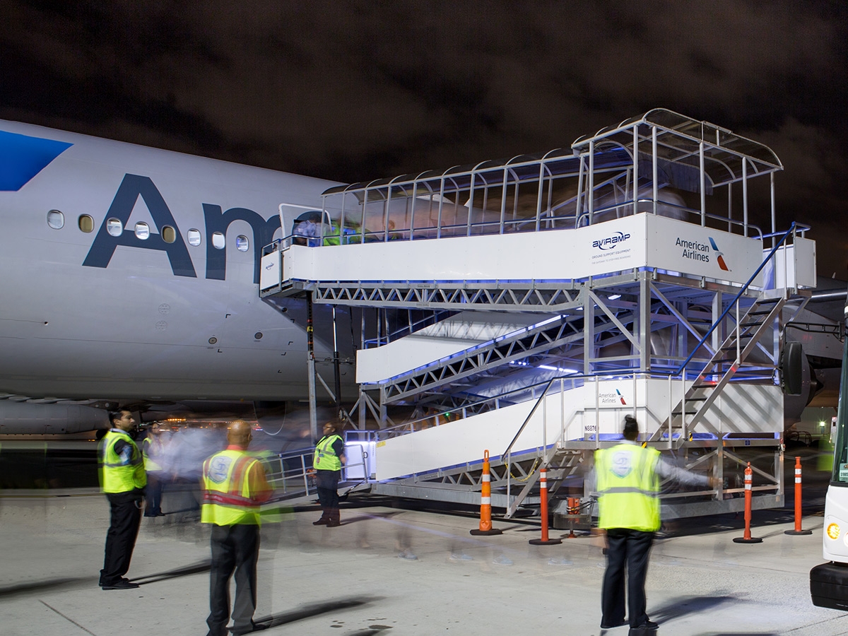 airport ground crew and personnel are gathered near an airplane. A mobile staircase is positioned at the aircraft's door, allowing passengers or crew to board or disembark. The scene likely takes place during the evening or night, highlighting airport activity and operational procedures during midnight hours.