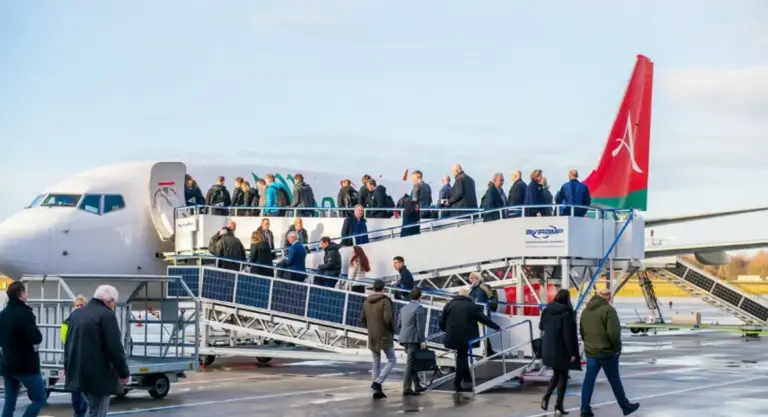 Passengers walking up the boarding stairs to enter the airplane at the airport, with some people waiting nearby, all under a partly cloudy sky.