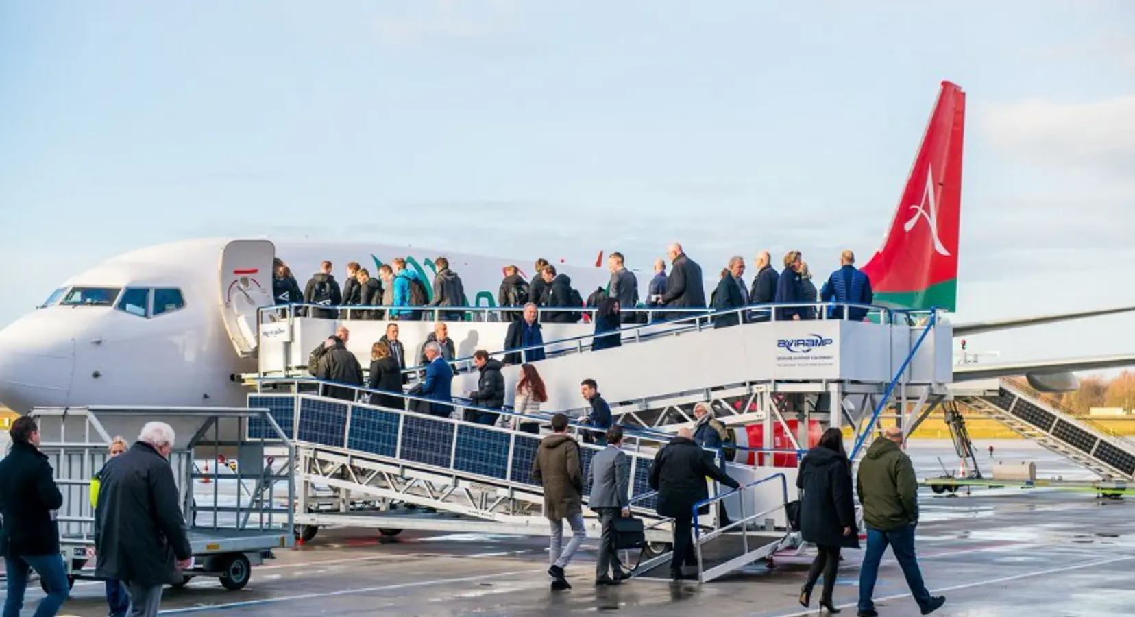 Passengers walking up the boarding stairs to enter the airplane at the airport, with some people waiting nearby, all under a partly cloudy sky.
