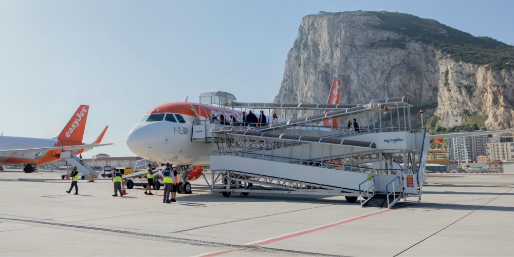 An easyJet plane is parked at the gate, with passengers boarding using a mobile staircase against a backdrop of mountains and a city skyline.