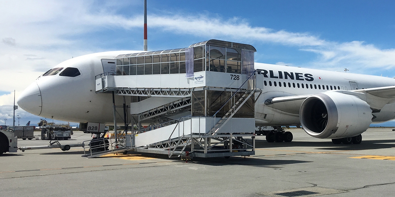 A plane is stationed at the terminal with a mobile Aviramp boarding ramp in place, as passengers prepare to board or exit under clear skies.