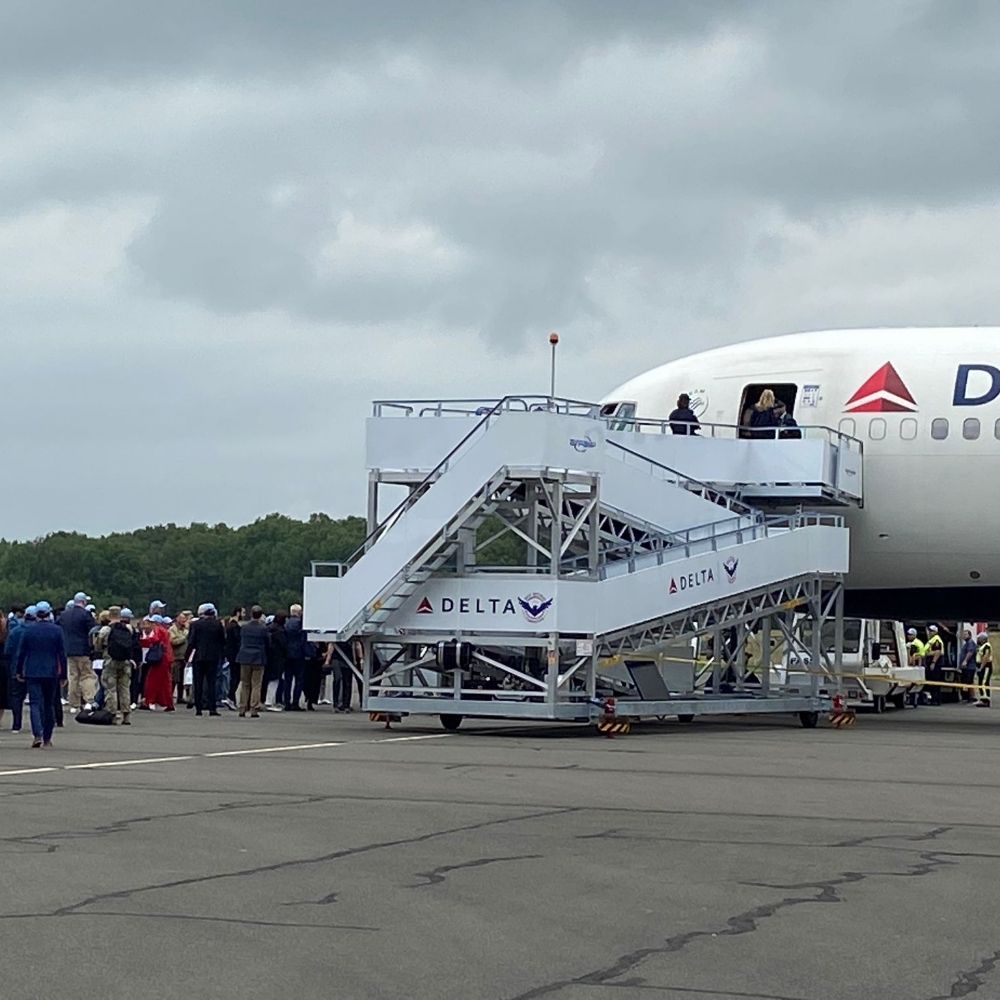 Travelers walking toward a Delta airplane as it prepares for departure, with ground crew and fellow passengers nearby against a backdrop of overcast skies.
