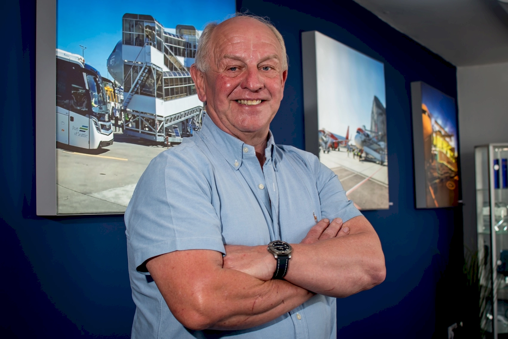Man in casual attire with arms crossed, standing in front of a wall decorated with aviation photographs, smiling confidently.