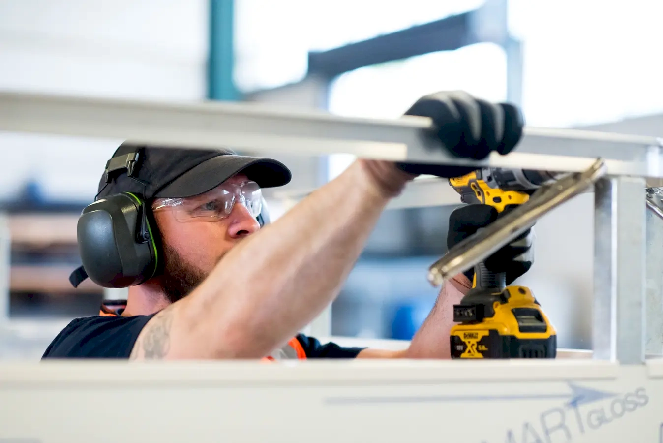 A worker engaged in construction or maintenance work within an industrial environment. Wearing essential safety gear—such as ear protection, safety glasses, gloves, and a work vest—the worker emphasizes safety and professionalism on the job site. Using a cordless drill, the worker appears to be assembling, securing, or repairing a metal structure, showcasing craftsmanship and attention to detail. The setting suggests a busy manufacturing plant, construction site, or factory where precision and safety are paramount. This image highlights the importance of safety protocols in industrial work, as well as the skill and focus required for tasks like assembly or maintenance. It also reflects the industrious spirit of workers who contribute to building and maintaining infrastructure, emphasizing the value of teamwork, safety, and technical expertise in industrial environments.