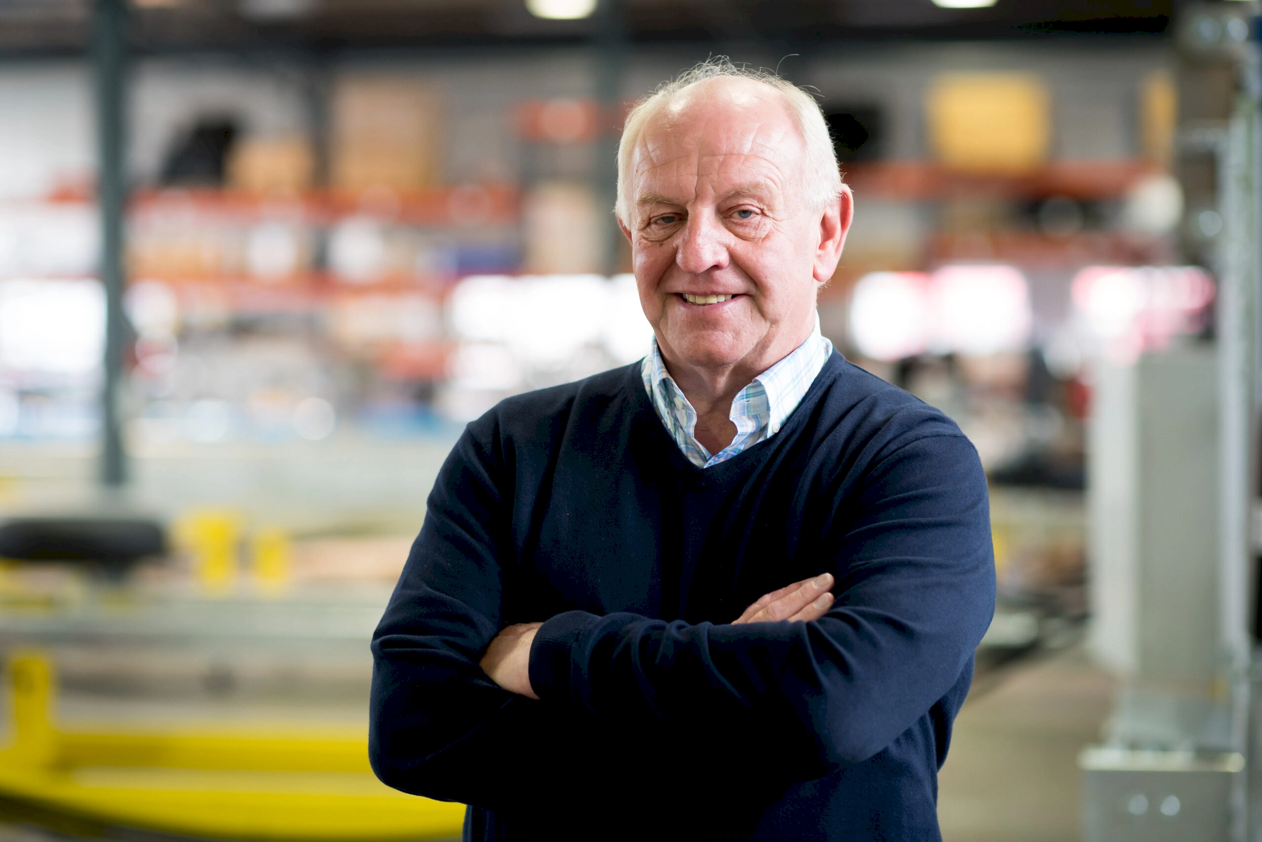 Graham Corfield, CEO of Aviramp wears a dark navy jumper over a light blue shirt, stands with his arms crossed and smiling in his Telford factory with shelving and storage visible in the background.