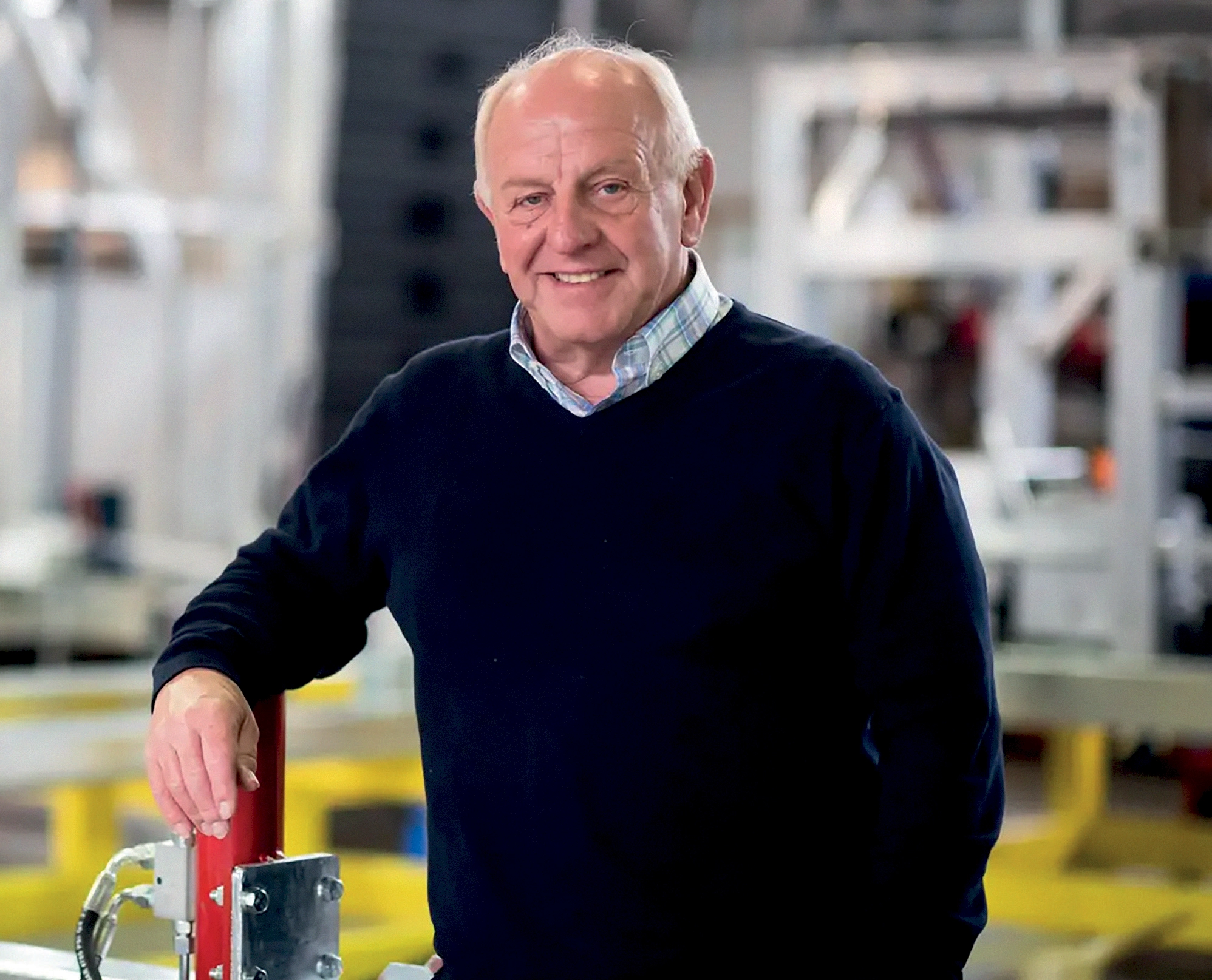 A a confident and approachable boss or manager in an industrial or manufacturing environment. His casual yet professional attire, combined with his relaxed posture and friendly smile, suggests that he is comfortable and confident in his leadership role. The background, featuring machinery and equipment, indicates that he works in a hands-on industry, possibly overseeing production, operations, or technical teams. His demeanour reflects qualities of a good leader—experienced, approachable, and supportive of his team. The setting highlights the importance of strong management in a busy work environment, ensuring that operations run smoothly while fostering a positive workplace culture. Overall, this image emphasizes the role of a leader who is knowledgeable, trustworthy, and committed to their work and team.