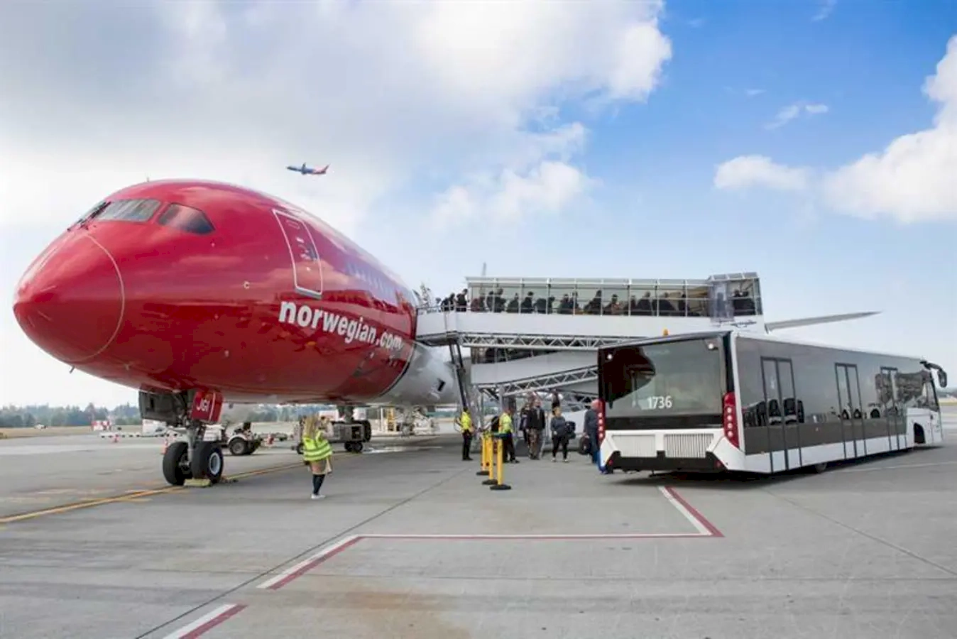 Norwegian Airlines aircraft parked at the airport, with a mobile staircase connected to it for passengers to board or leave, and ground staff working nearby under a partly cloudy sky.