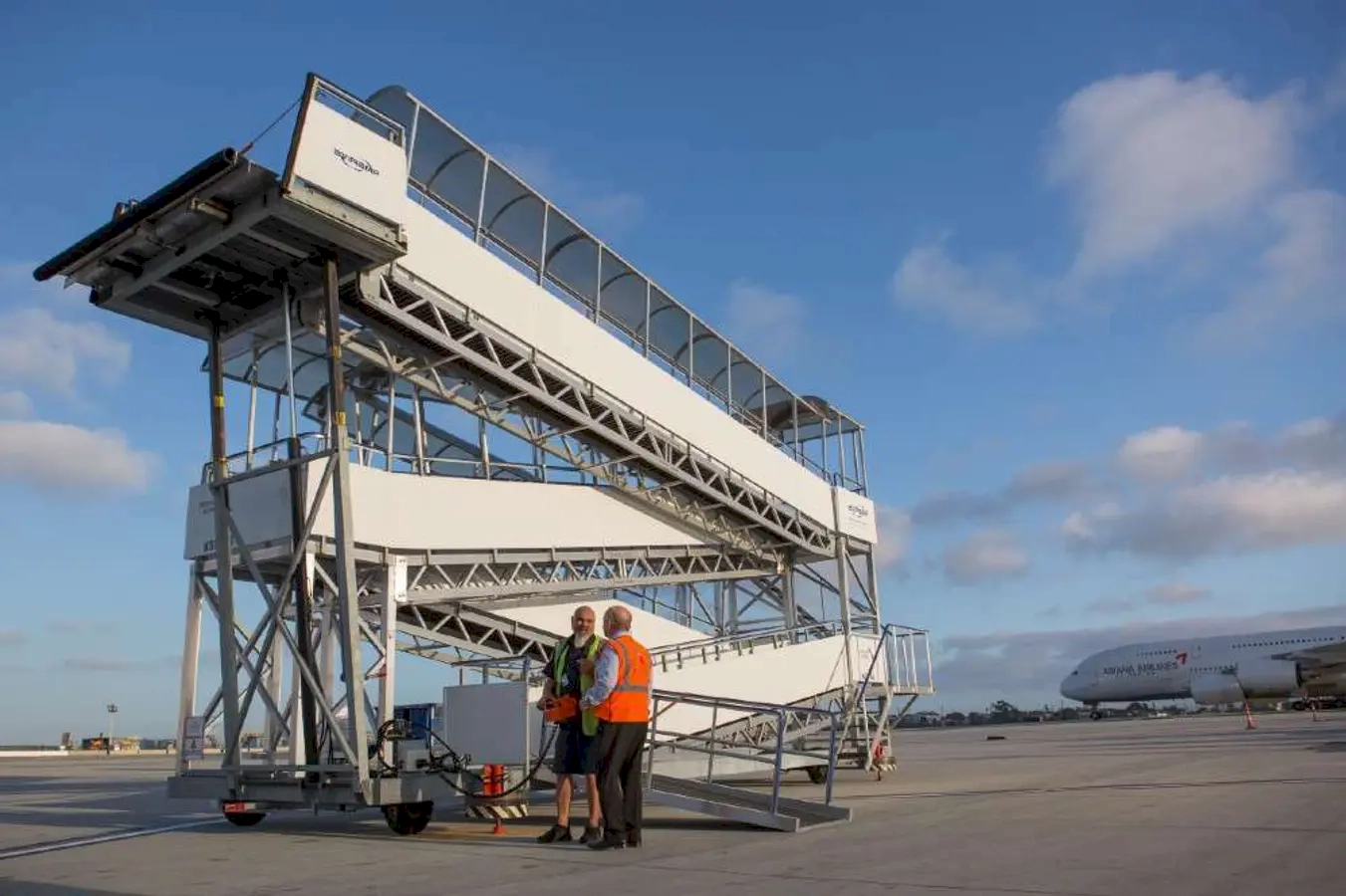 Two airport ground staff in orange safety vests standing and talking near a mobile staircase, with a large airplane parked nearby under a partly cloudy sky.