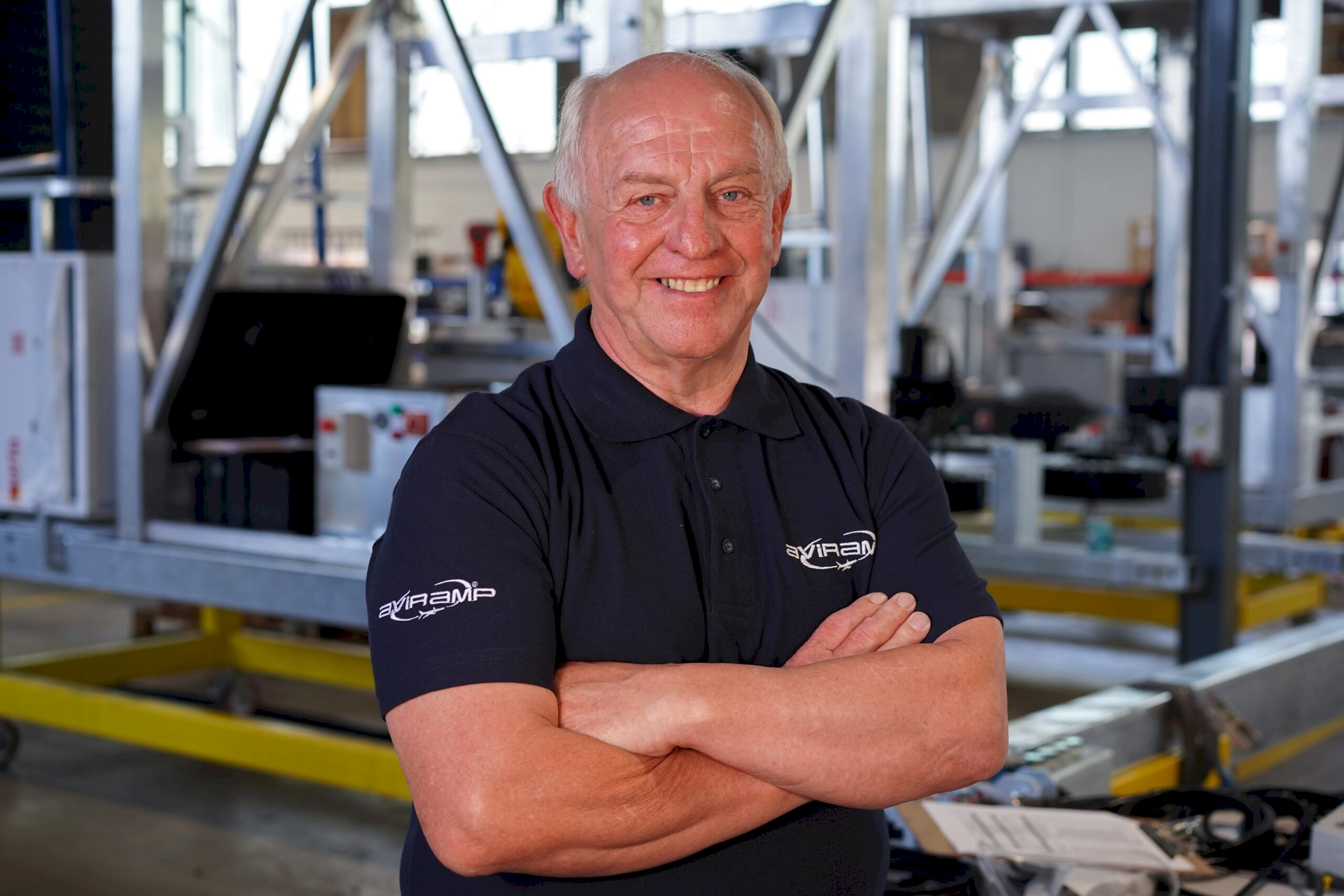 A cheerful man posing confidently with his arms crossed amidst the busy environment of an industrial workspace.