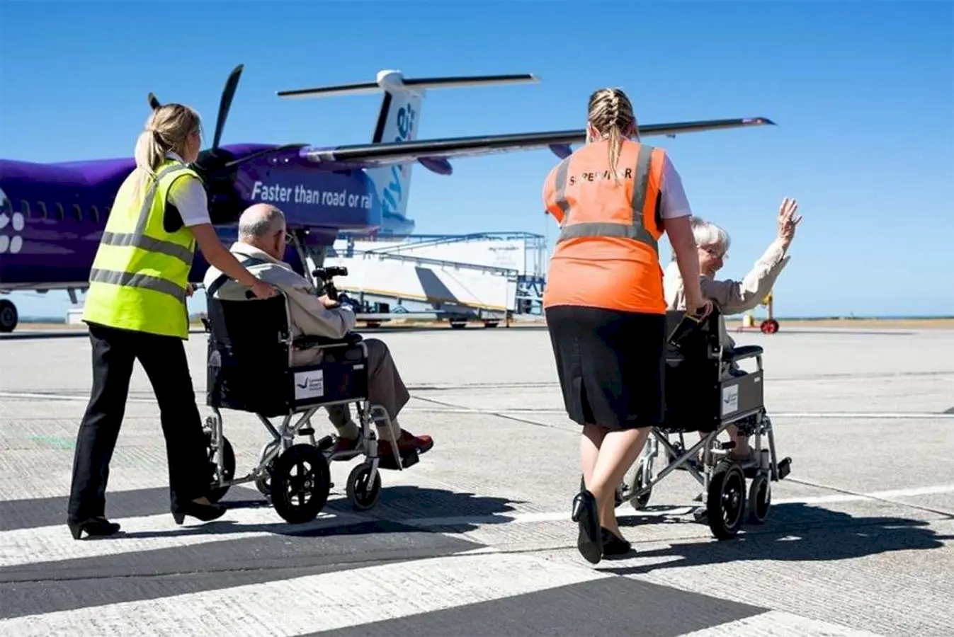 Airport staff helping passengers in wheelchairs to board or disembark from an aircraft on the tarmac, with a small airplane visible in the background under a clear blue sky.