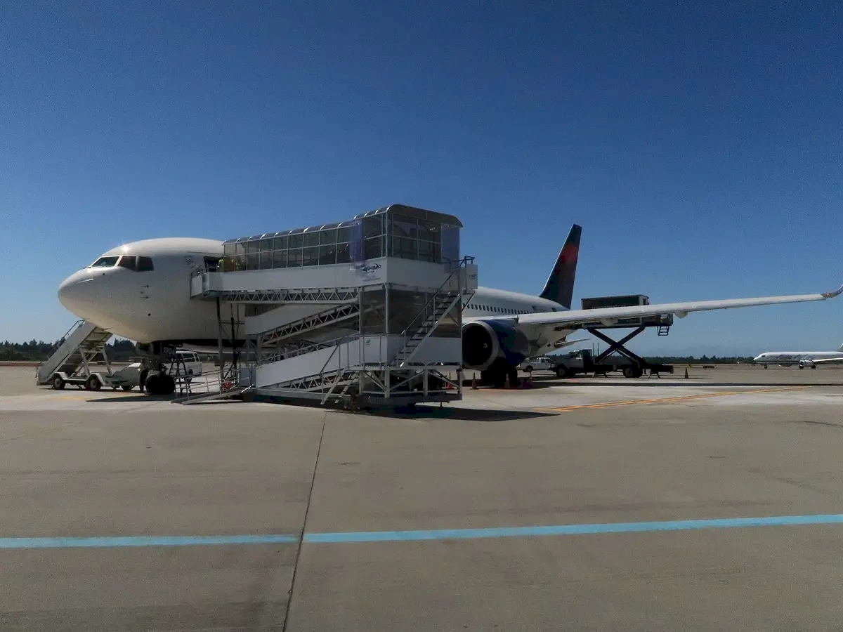 A moment of transition in air travel, where an airplane is parked on the tarmac, ready to welcome passengers. The boarding stairs positioned at the aircraft's door serve as a vital link between the ground crew and travelers, facilitating a smooth boarding process. The clear blue sky and the distant landscape—possibly mountains or open fields—add a sense of serenity and adventure, emphasizing the excitement of upcoming journeys. The bright sunlight and open space evoke feelings of optimism, exploration, and the thrill of discovering new destinations. This setup underscores the efficiency and organization behind commercial flights, showcasing the teamwork and planning that go into making air travel safe and comfortable. Whether it's a departure or arrival scene, it captures the anticipation and promise of travel, inviting viewers to imagine themselves embarking on an exciting trip to a distant place.