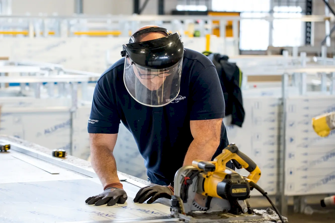 A muscular man engaged in a demanding task within an industrial or construction setting. His well-built physique suggests he is accustomed to physically intense work, emphasizing both power and endurance. Wearing essential safety equipment—such as gloves and a face shield—he demonstrates a commitment to safety while handling heavy-duty tools like a power saw. The focus and precision on his face indicate his experience and concentration, ensuring the task is completed efficiently and safely. The industrial backdrop, with tools and materials around him, underscores the environment where strength and technical skill are vital. This image celebrates craftsmanship, physical fitness, and the importance of safety protocols in manual labour jobs. Overall, it portrays a hardworking individual using his strength and expertise to accomplish a challenging task, embodying resilience, skill, and dedication in a tough working environment.