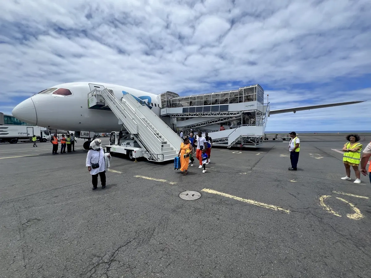 Passengers leaving the airplane using mobile stairs at the airport, with some carrying luggage and others waiting nearby.