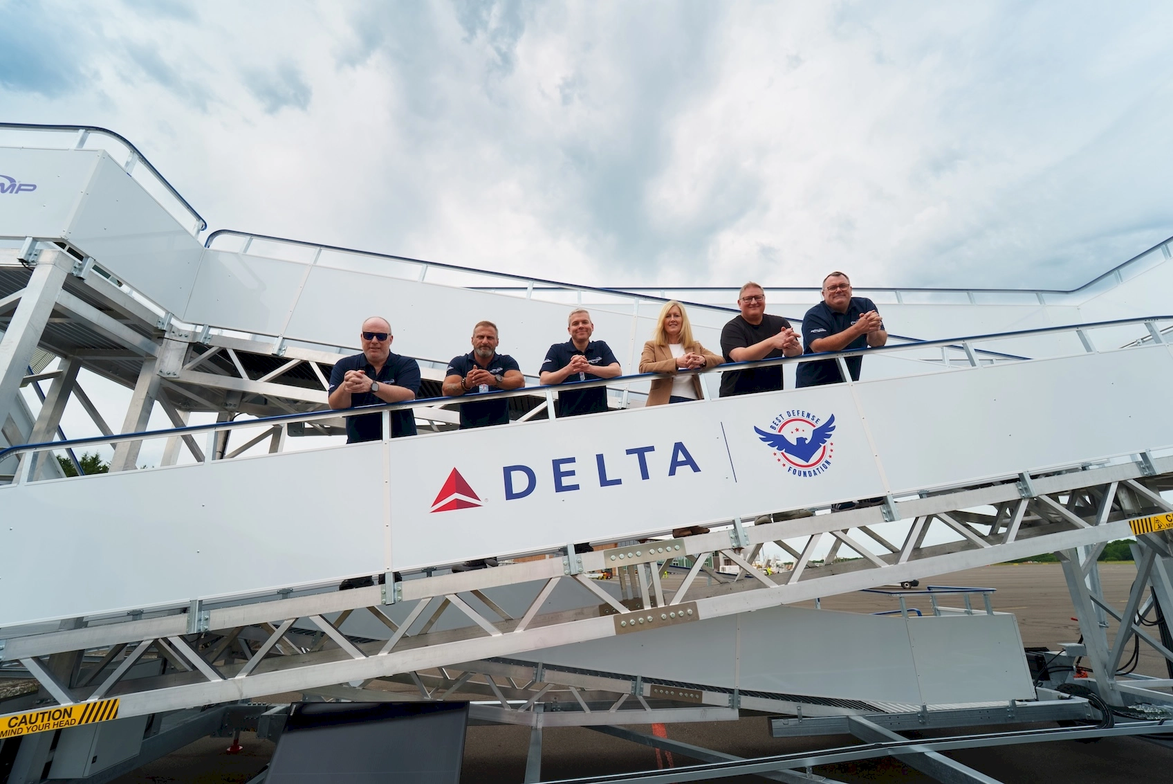 A group of six people pose happily on an outdoor airport staircase featuring Delta and Best Defence Foundation logos, with a clear sky in the background.