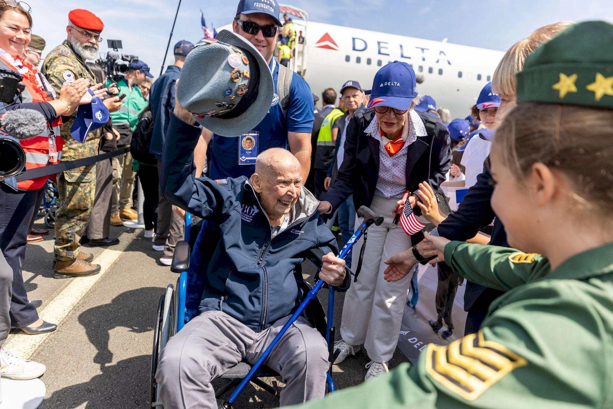 A veteran in a wheelchair is warmly greeted with cheers and applause, waving his hat in celebration amidst supporters and military personnel at the airport.