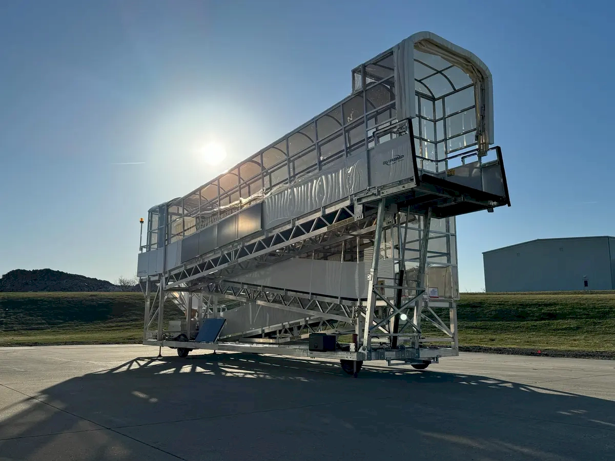 A moment of anticipation and readiness at an airport, where the mobile aircraft boarding stairs stand prepared on the tarmac. The bright sunshine and clear blue sky create a vibrant, inviting atmosphere, emphasizing the excitement of travel and adventure that lies ahead. The stairs, likely made of lightweight yet sturdy materials, are ready to connect passengers from the ground to the aircraft door, symbolizing the beginning of a journey. The sunlight casting shadows adds depth and dimension to the scene, highlighting the sleek design and mobility of the boarding equipment.