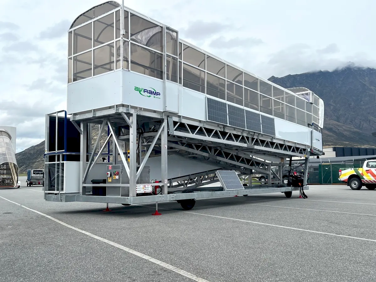 This scene captures a sleek, modern aircraft boarding setup that highlights the advancements in airport infrastructure. The mobile staircase, designed with contemporary materials and possibly featuring integrated lighting or branding, underscores the emphasis on passenger convenience and safety. Set against a stunning natural backdrop of mountains and a partly cloudy sky, the scene evokes a sense of adventure and exploration. The positioning of the staircase on the tarmac suggests readiness for departure or arrival, symbolizing the seamless connection between travelers and their destinations. This setup not only facilitates efficient boarding and disembarking but also reflects the innovation within the aviation industry aimed at enhancing the overall travel experience. The combination of natural beauty and cutting-edge airport technology creates an inspiring image of how modern travel harmonizes with scenic landscapes, inviting travellers to embark on new journeys filled with discovery and excitement.