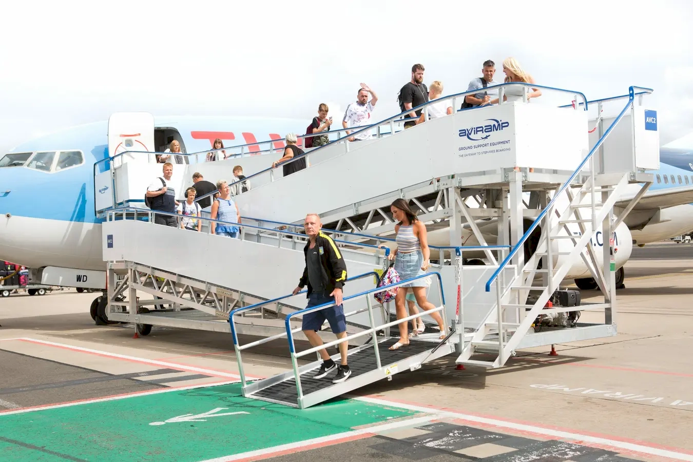 Passengers getting off an aircraft using mobile stairs at the airport, with some walking down the ramp and others waiting, all under a partly cloudy sky.