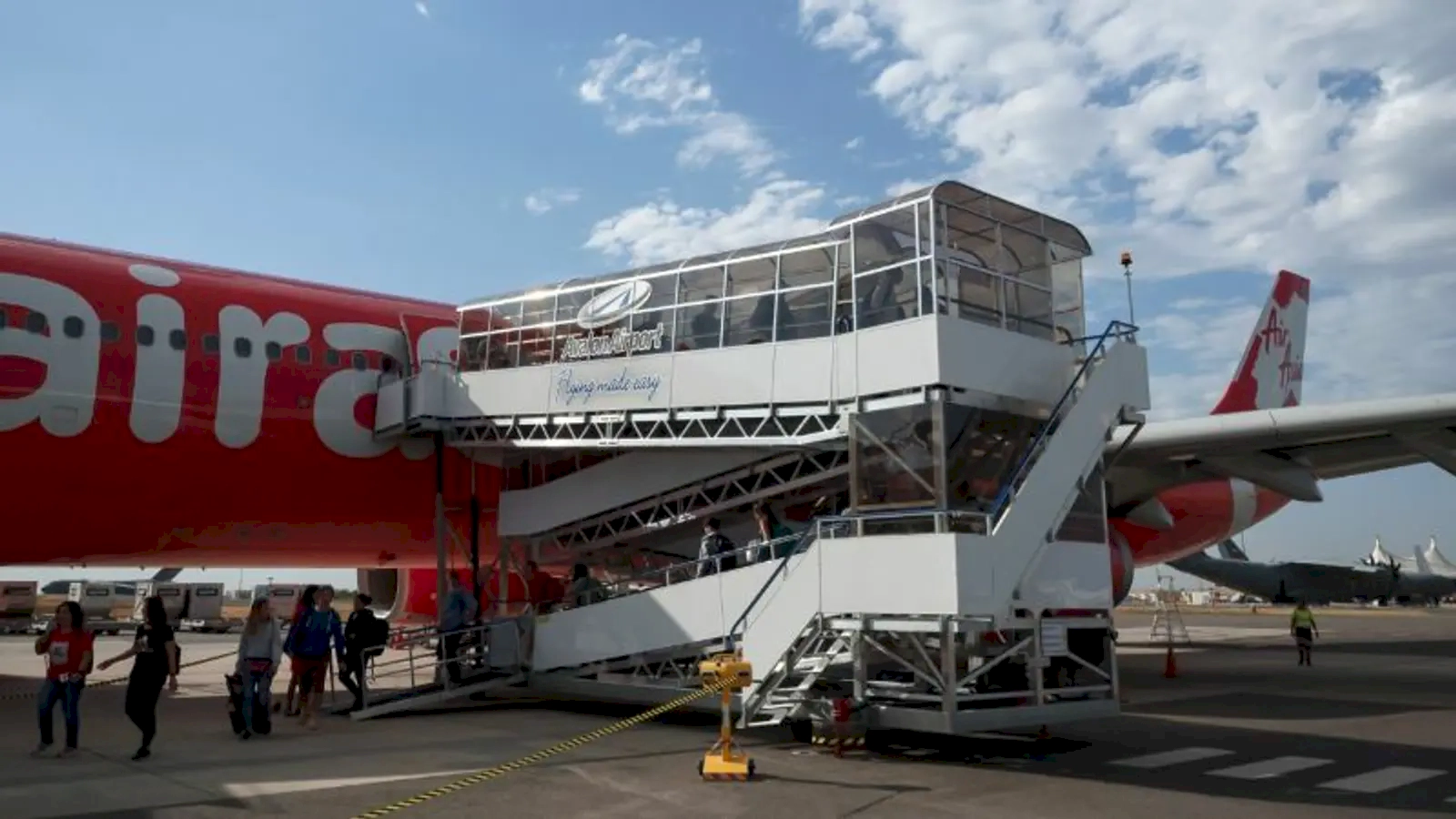 Passengers boarding an AirAsia airplane using a mobile staircase at the airport, with a clear sky above and other aircraft visible in the background.