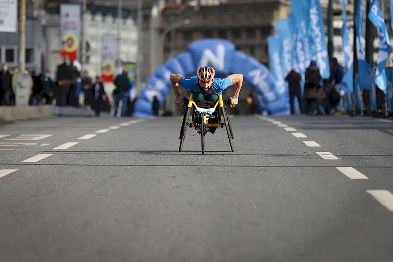 The inspiring spirit of perseverance and determination, capturing a wheelchair athlete during a competitive race or a community event. The athlete, dressed in a helmet and sports gear, exemplifies strength, focus, and resilience as they push forward on the track or city street. The presence of spectators and flags in the background indicates a lively, supportive environment, celebrating inclusivity and sportsmanship. This moment emphasizes the importance of accessibility, showcasing how adaptive sports enable individuals with disabilities to challenge themselves and achieve their goals. The urban setting adds a dynamic backdrop, symbolizing the breaking down of barriers and the integration of adaptive sports into everyday life. Overall, the image celebrates human spirit, courage, and the power of sports to inspire and unite people across communities.